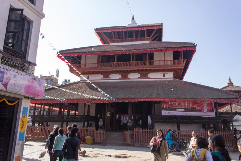A multi-tiered traditional building with a red roof at a bustling square, with prayer flags, people walking, and sitting on steps, under a clear blue sky.