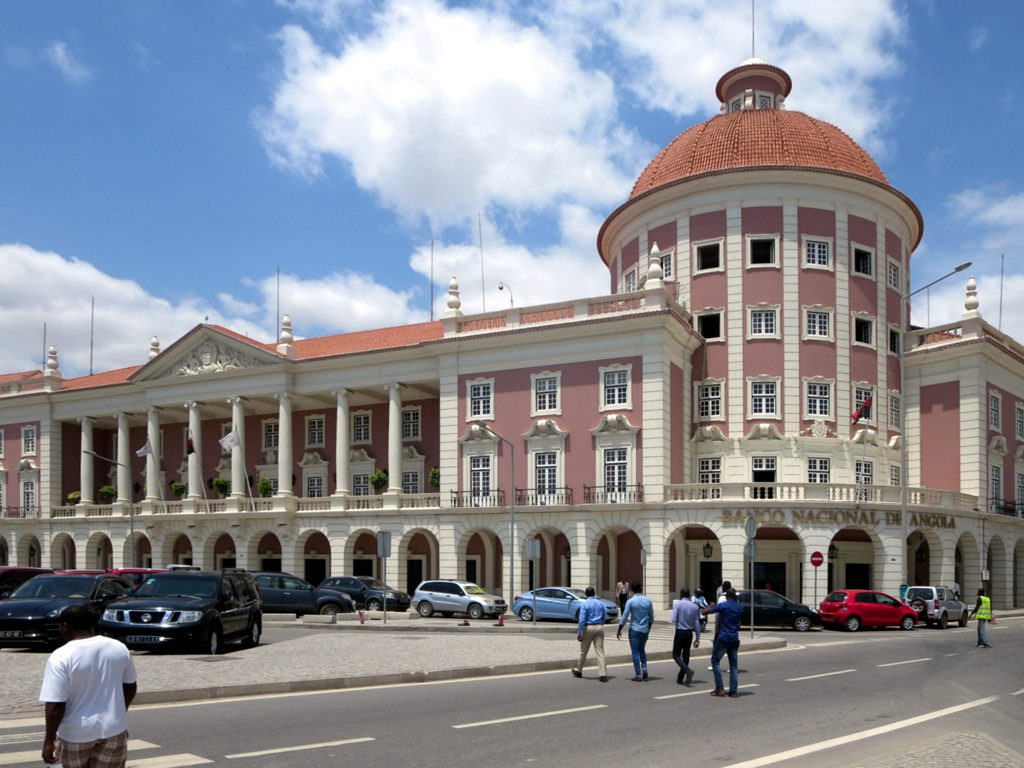 A large colonial-style pink building with white trim and a red-tiled dome, identified as the "Banco Nacional de Angola" on the facade, with pedestrians and parked cars in front.