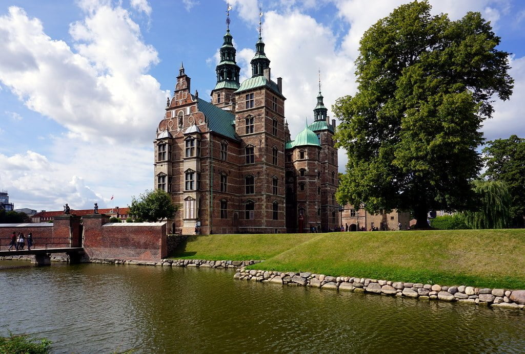 Alt text: A historic red brick castle with green copper spires overlooking a tranquil moat, surrounded by manicured lawns and a large tree under a partly cloudy sky.