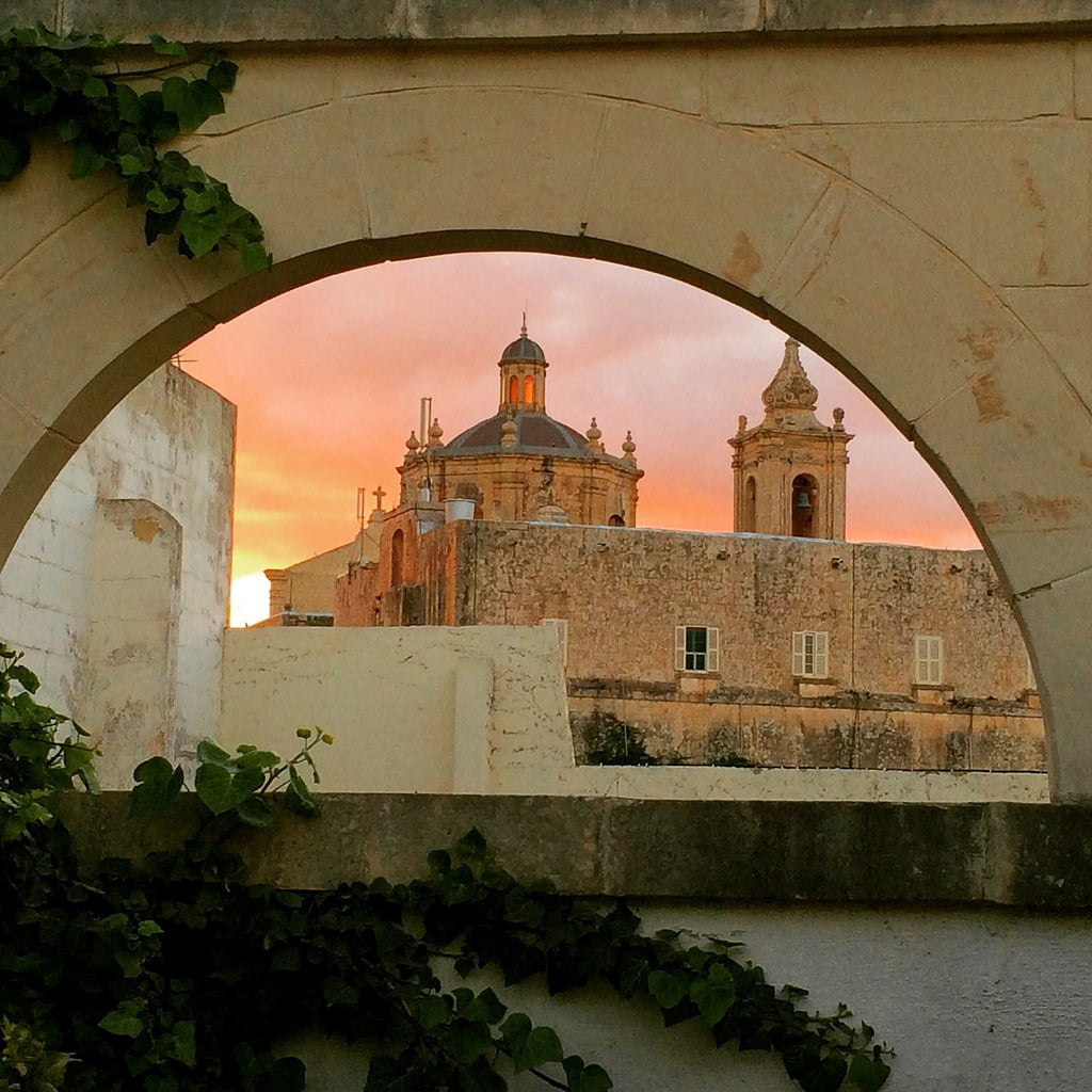 An old stone church with a dome and bell tower seen through an archway during sunset, with the sky glowing in hues of pink and orange. Ivy creeps up one side of the arch.