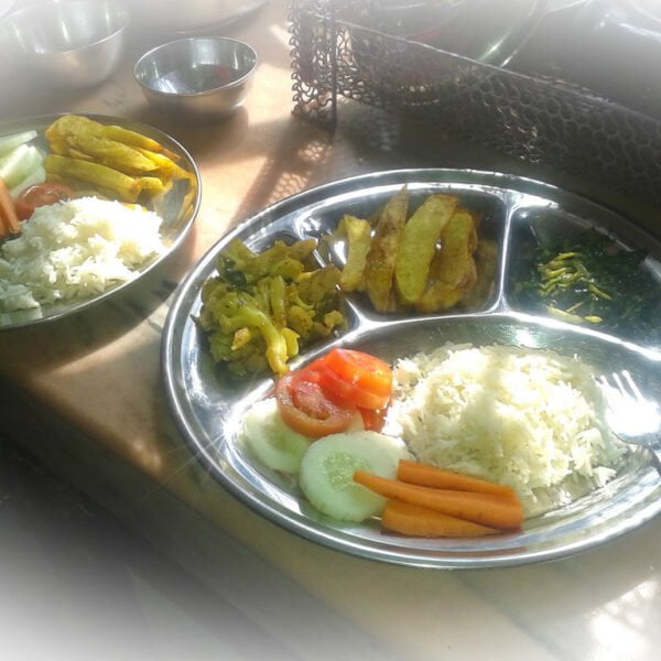 A plated meal consisting of white rice, sautéed vegetables, slices of tomato and cucumber, and fried plantains, with a fork on the side, illuminated by natural light.