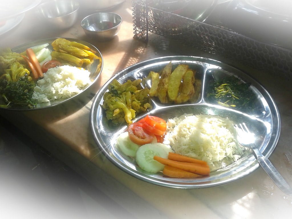 A plated meal consisting of white rice, sautéed vegetables, slices of tomato and cucumber, and fried plantains, with a fork on the side, illuminated by natural light.
