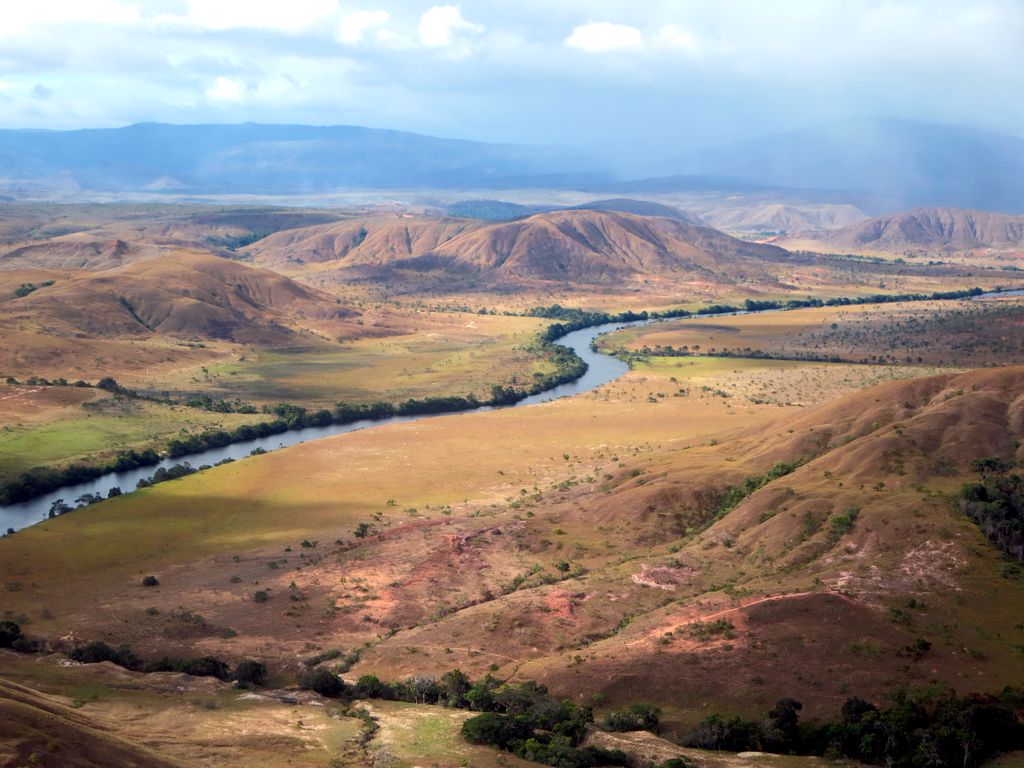 Aerial view of a meandering river cutting through a dry, hilly landscape with sparse vegetation and a backdrop of mountains partially obscured by haze.