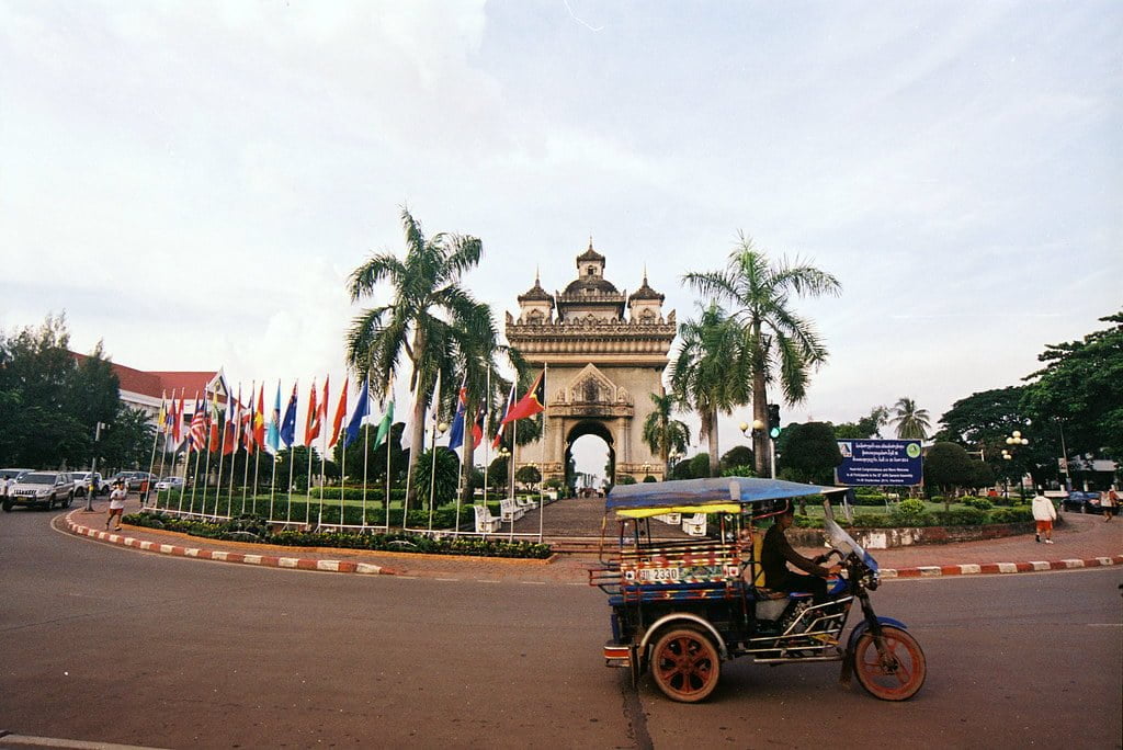 A tuk-tuk in the foreground on the right with a driver, in front of a row of international flags and a monumental gate with arches and multiple tiers, possibly in a Southeast Asian country, under a blue sky with scattered clouds.