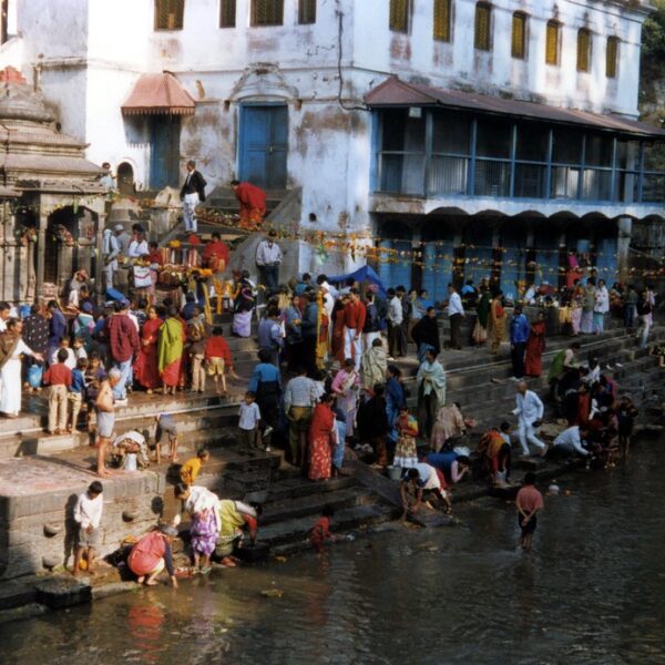 A bustling scene at a riverside with people engaging in various activities on the steps, such as washing, socializing, and walking, against the backdrop of traditional buildings and a temple entrance.
