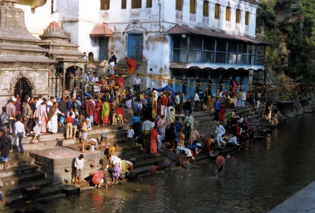 A bustling scene at a riverside with people engaging in various activities on the steps, such as washing, socializing, and walking, against the backdrop of traditional buildings and a temple entrance.