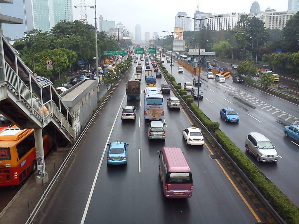 Alt text: A busy city street with multiple lanes of traffic, including cars, buses, and motorcycles, with an overhead pedestrian walkway and urban buildings in the background on a cloudy day.