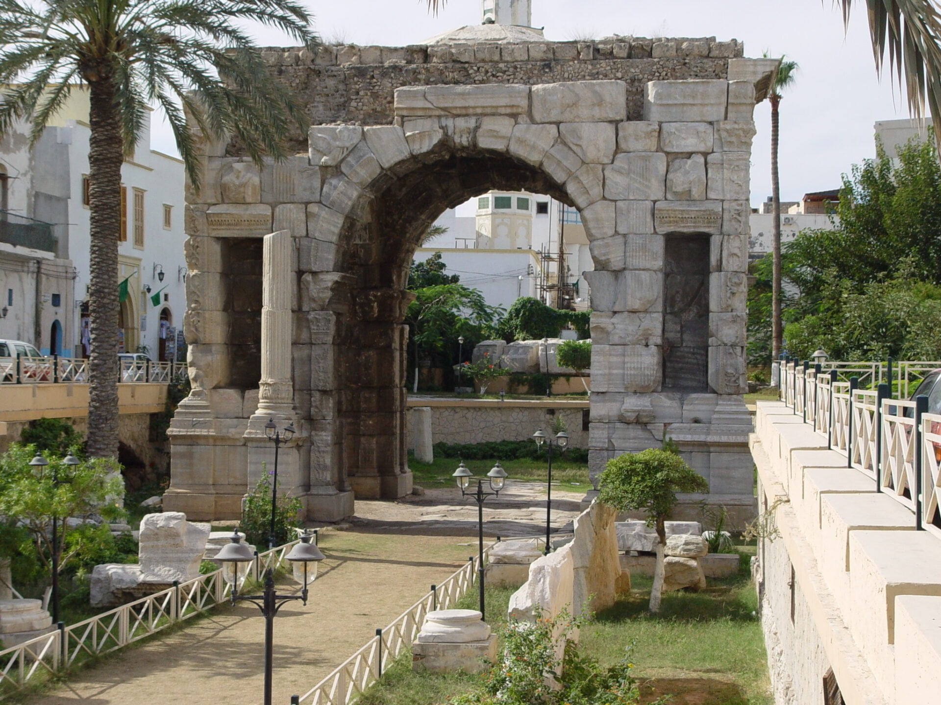 An ancient Roman triumphal arch constructed from large stone blocks, surrounded by palm trees, street lamps, and a sidewalk, with modern buildings in the background.