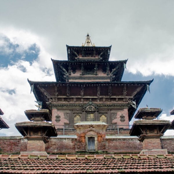 A multi-tiered pagoda-style temple with intricate wood carvings and a red clay tile roof against a backdrop of a cloudy sky.