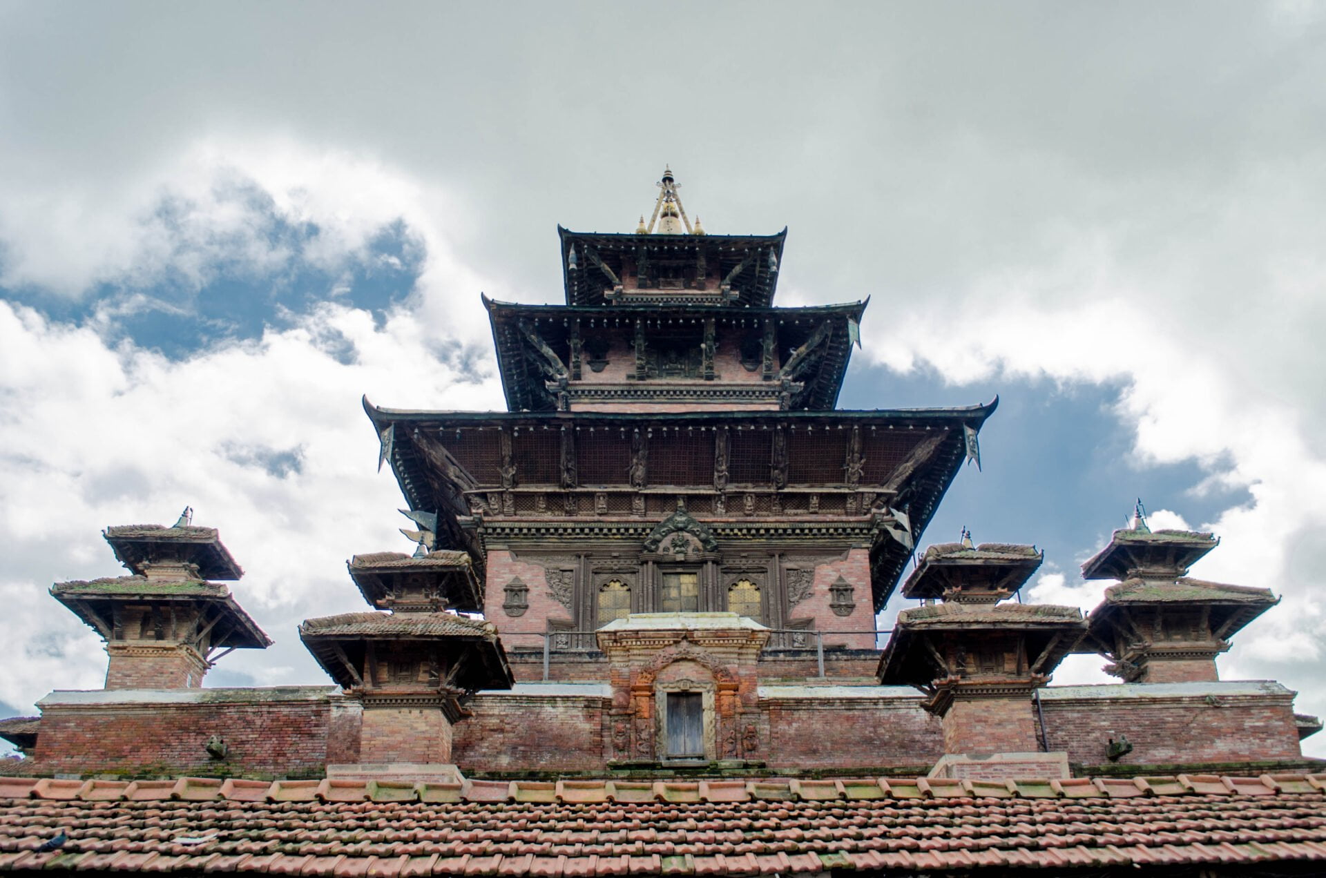 A multi-tiered pagoda-style temple with intricate wood carvings and a red clay tile roof against a backdrop of a cloudy sky.