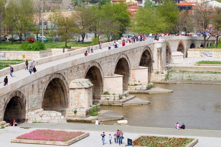 An ancient stone bridge with multiple arches spans a river, populated with pedestrians, in a lush urban park setting.