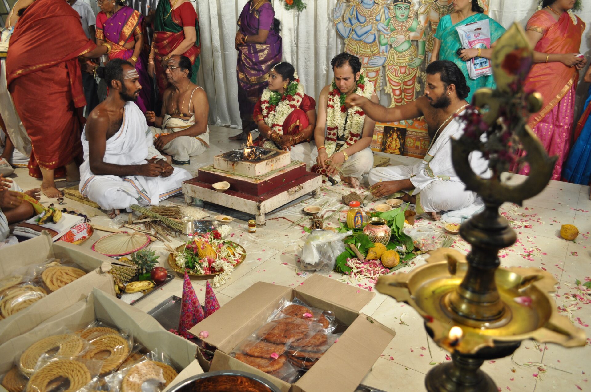 A traditional Hindu wedding ceremony with a bride and groom sitting by a sacred fire, surrounded by offerings and guests, with a priest conducting rituals.