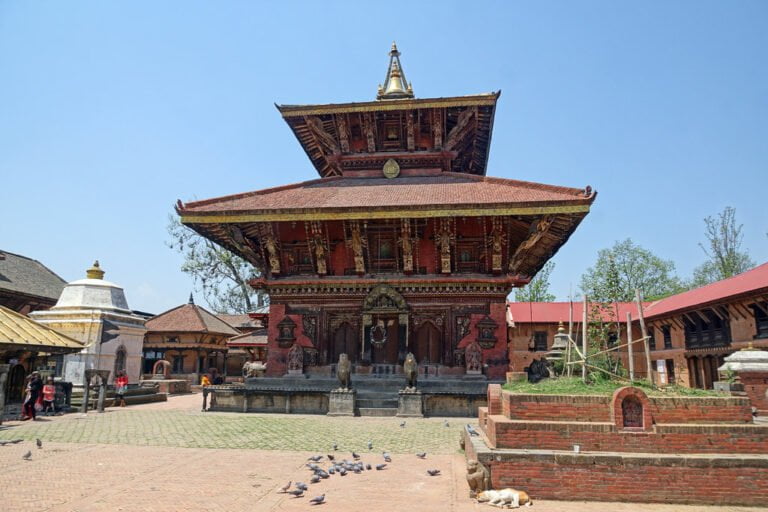 Alt text: A traditional Nepalese temple with intricate wood carvings and a tiered pagoda-style roof, located in a square with scattered pigeons and a few people walking around.