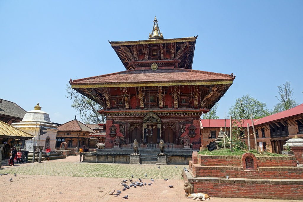 Alt text: A traditional Nepalese temple with intricate wood carvings and a tiered pagoda-style roof, located in a square with scattered pigeons and a few people walking around.