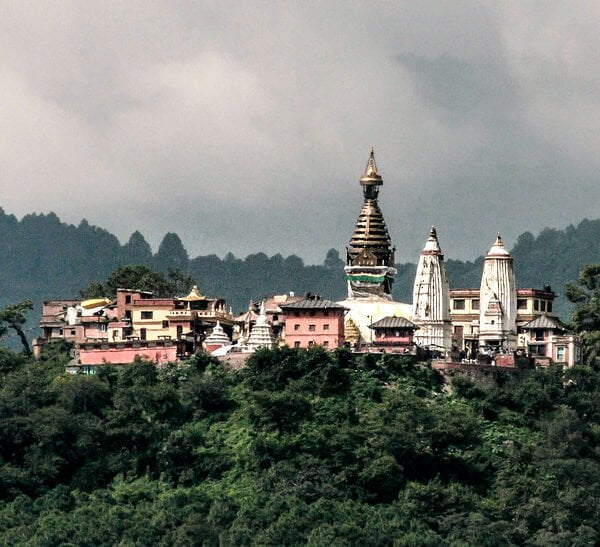 Colorful buildings and a temple with multiple spires nestled on a densely forested hill with a cloudy sky in the background.