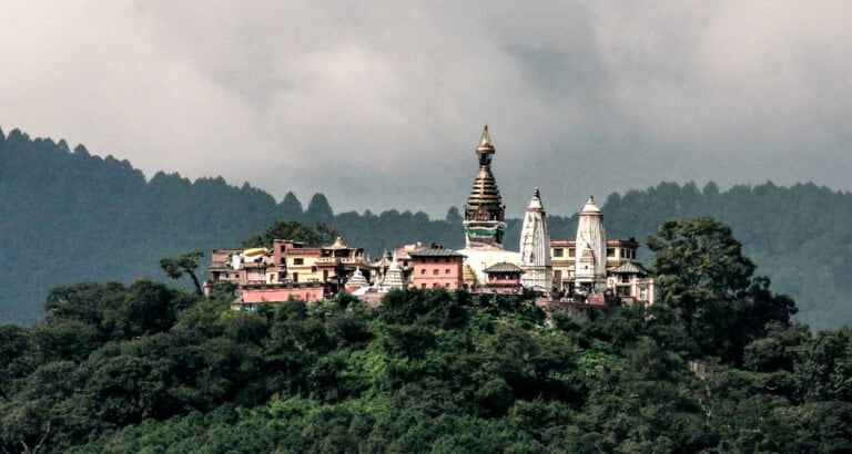 Colorful buildings and a temple with multiple spires nestled on a densely forested hill with a cloudy sky in the background.