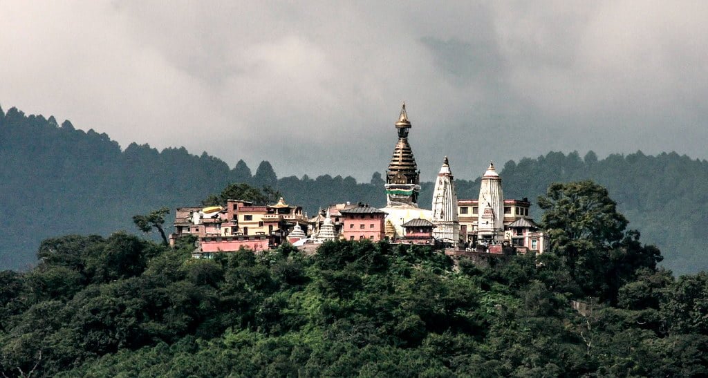 Colorful buildings and a temple with multiple spires nestled on a densely forested hill with a cloudy sky in the background.