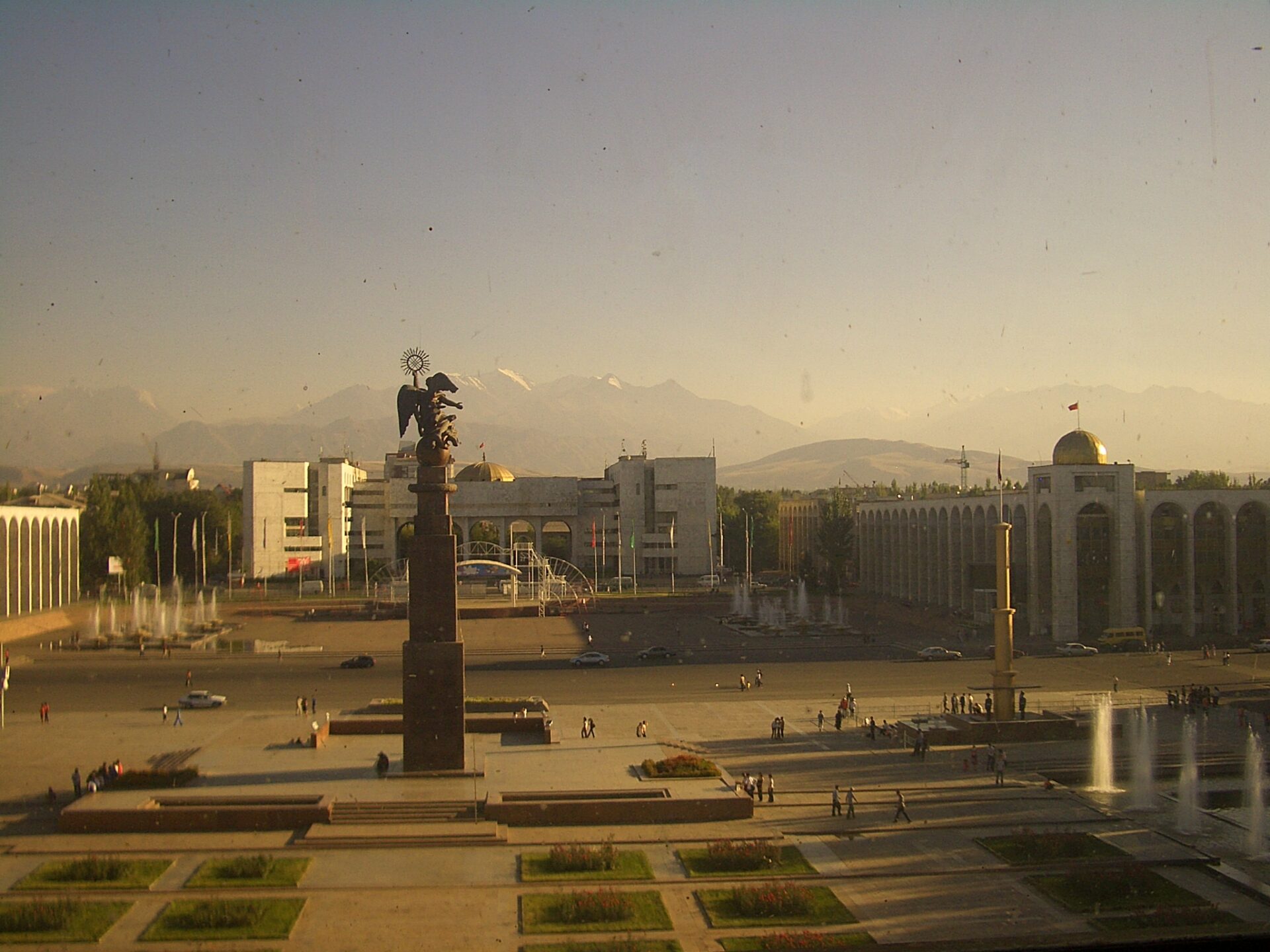 A view of a city square with a monument in the center, people walking, buildings in the background, and mountains on the horizon, viewed through a window with reflections and spots.