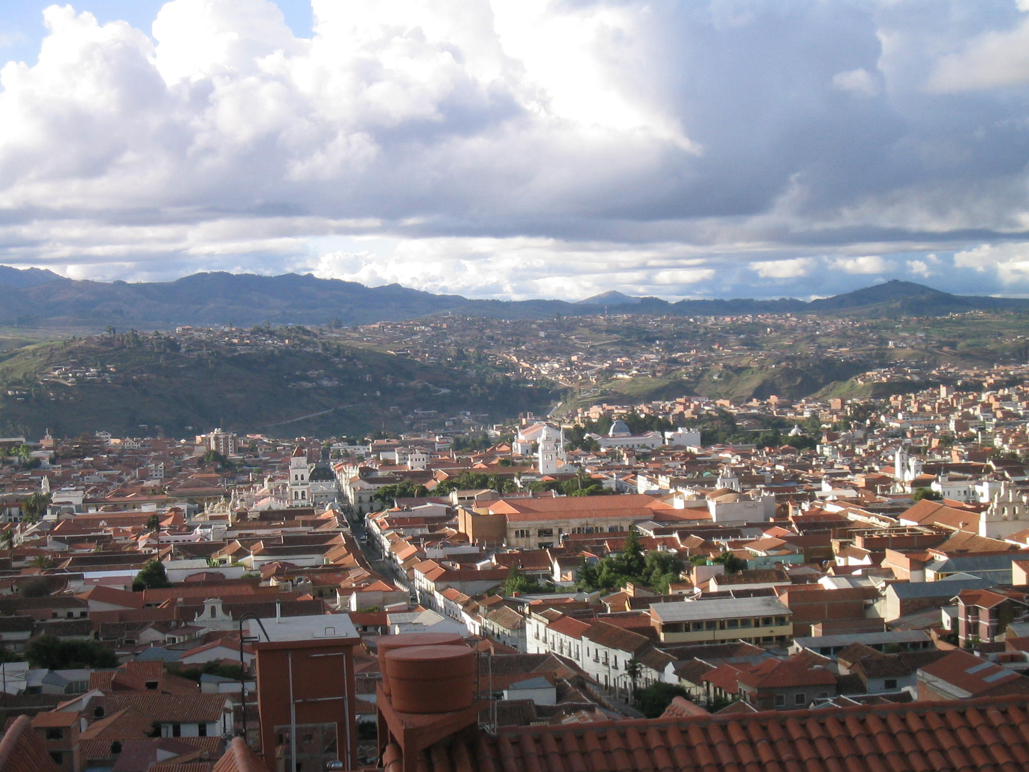 A panoramic view of a densely built city with terra-cotta rooftops, nestled in a valley with rolling hills in the background under a partly cloudy sky.