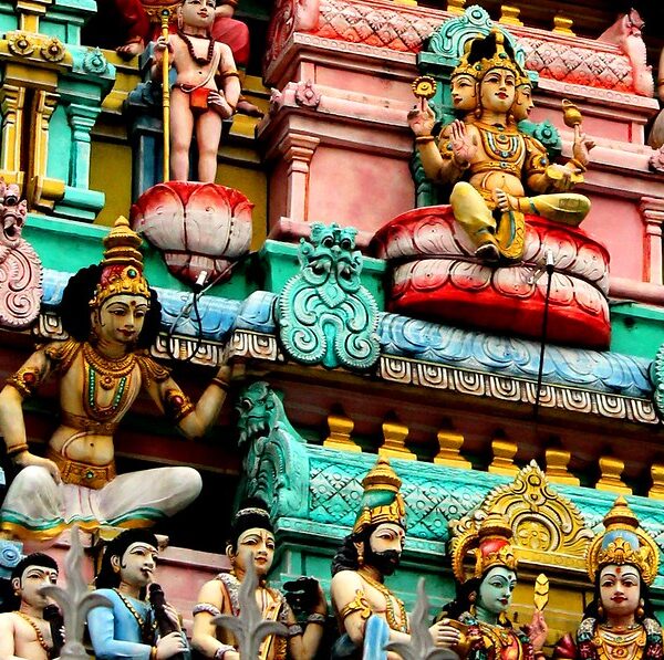 Close-up of a colorful Hindu temple façade featuring an intricate array of sculpted figures and deities with ornate detailing in vibrant hues of pink, blue, green, and gold.