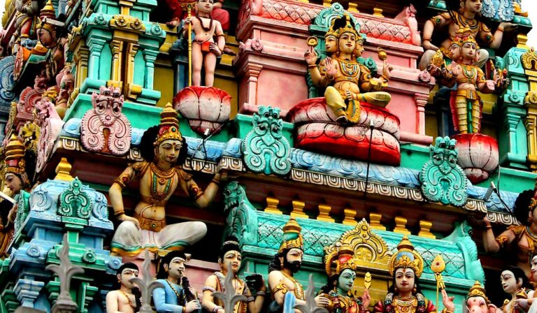 Close-up of a colorful Hindu temple façade featuring an intricate array of sculpted figures and deities with ornate detailing in vibrant hues of pink, blue, green, and gold.