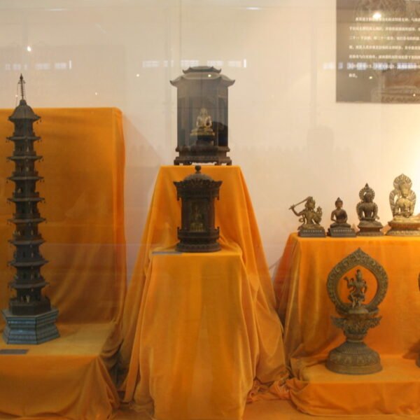 A display of intricate Asian artifacts, including a tall pagoda structure and various statues of deities and mythical creatures, presented on orange cloths behind a glass enclosure.