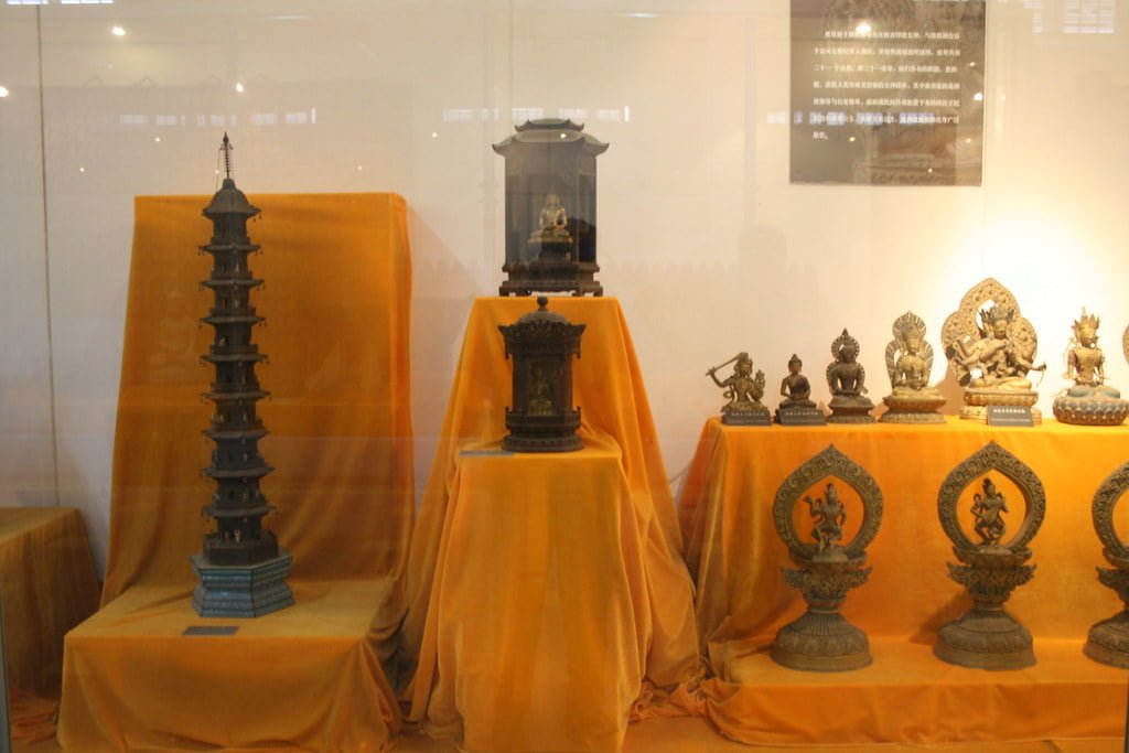 A display of intricate Asian artifacts, including a tall pagoda structure and various statues of deities and mythical creatures, presented on orange cloths behind a glass enclosure.