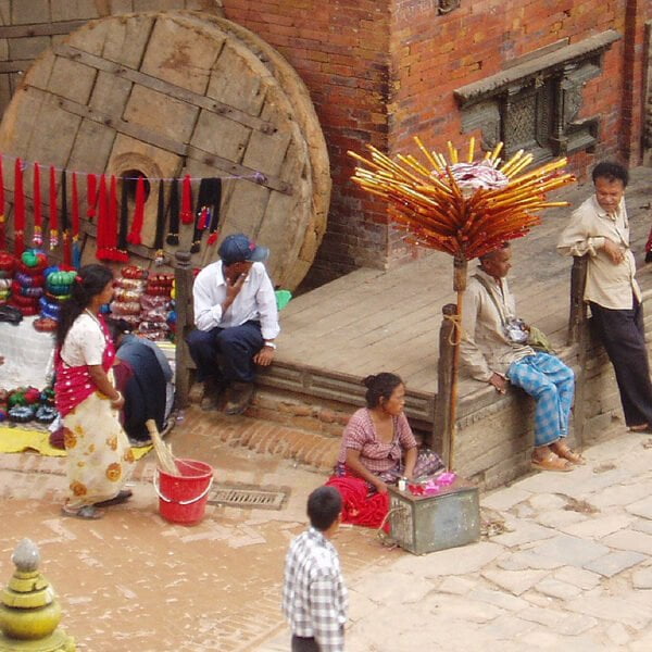 Alt text: A street scene with various people engaged in daily activities: some sitting and talking, others selling colorful goods on the pavement next to large wooden wheels, with traditional architecture in the background. An eye-catching display featuring numerous orange sticks with tips facing upwards dominates the foreground.