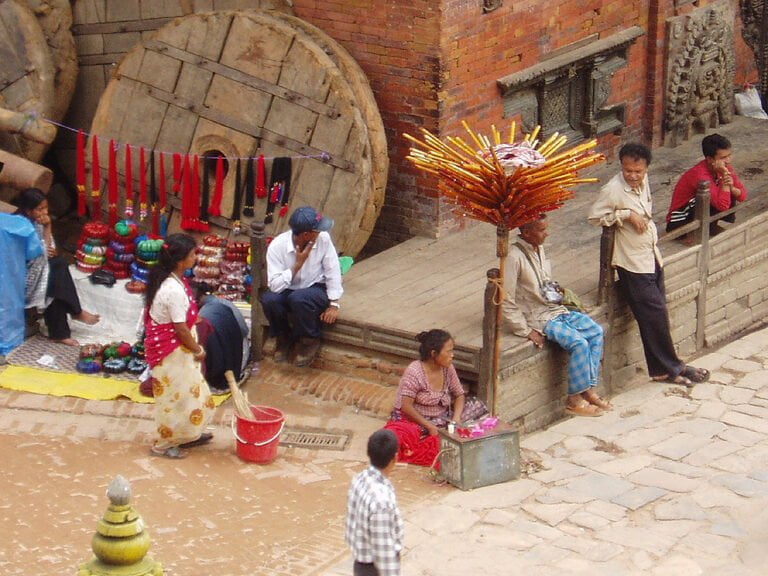 Alt text: A street scene with various people engaged in daily activities: some sitting and talking, others selling colorful goods on the pavement next to large wooden wheels, with traditional architecture in the background. An eye-catching display featuring numerous orange sticks with tips facing upwards dominates the foreground.