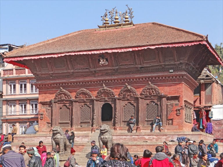 An ornate red-brick temple adorned with wood carvings and statues, flanked by large stone lion sculptures, amidst a bustling crowd of people in a square.