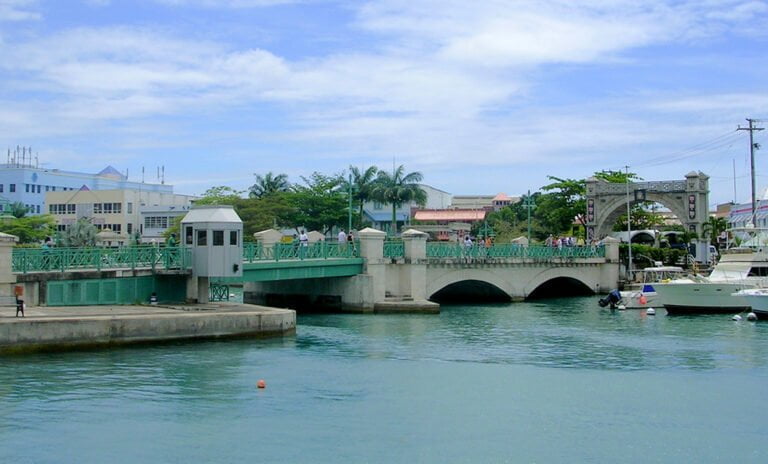 A scenic view of a green-painted bridge over calm blue waters with boats docked nearby and tropical architecture in the background.