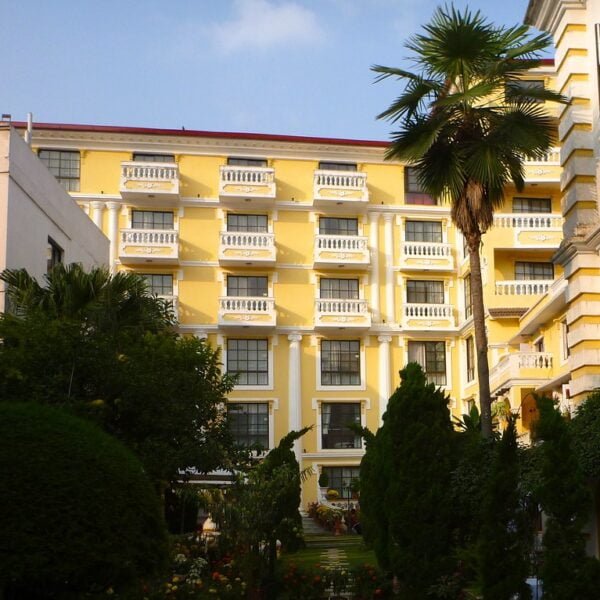 A yellow multi-story building with white balconies, surrounded by lush greenery and palm trees, under a clear sky.