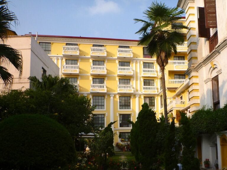 A yellow multi-story building with white balconies, surrounded by lush greenery and palm trees, under a clear sky.