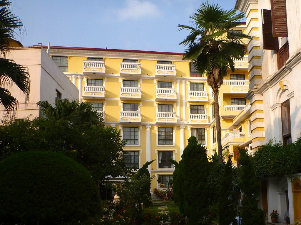 A yellow multi-story building with white balconies, surrounded by lush greenery and palm trees, under a clear sky.