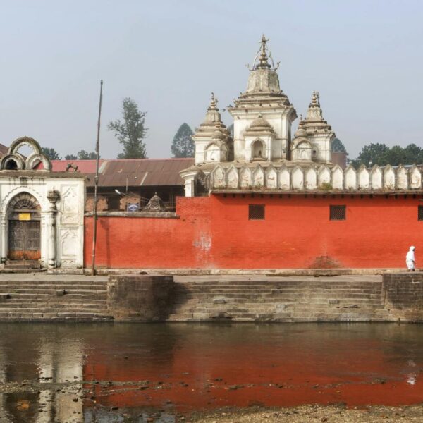 A Nepalese temple with a bright red wall, white decorative religious structures, and stepped ghats leading to a river. A solitary figure dressed in white walks along the riverbank.