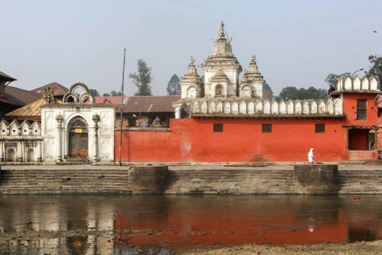 A Nepalese temple with a bright red wall, white decorative religious structures, and stepped ghats leading to a river. A solitary figure dressed in white walks along the riverbank.