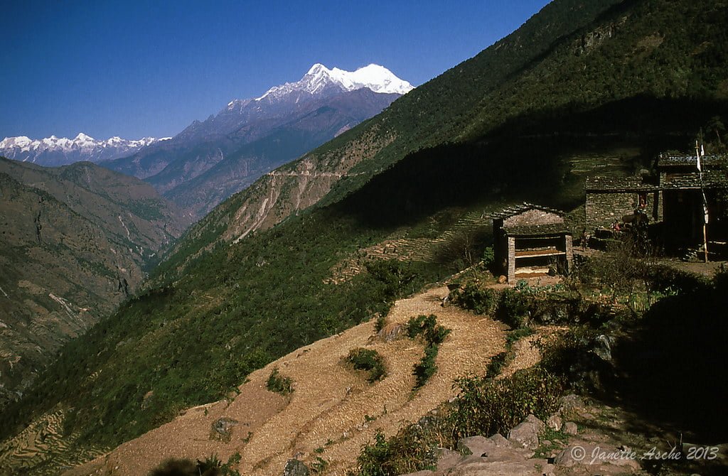 A panoramic view of terraced fields and traditional stone structures in the foreground with the snow-capped Himalayan mountain range in the background under a clear blue sky.