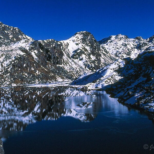 A serene alpine landscape featuring a clear, calm lake reflecting surrounding snow-dusted mountains under a deep blue sky.