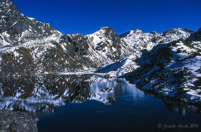 A serene alpine landscape featuring a clear, calm lake reflecting surrounding snow-dusted mountains under a deep blue sky.