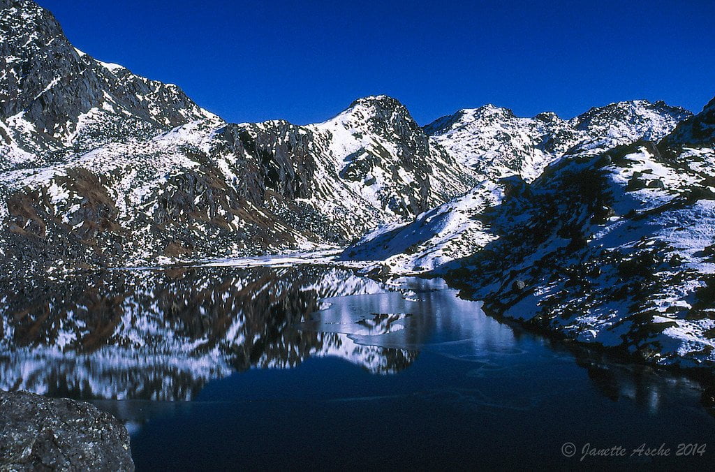 A serene alpine landscape featuring a clear, calm lake reflecting surrounding snow-dusted mountains under a deep blue sky.