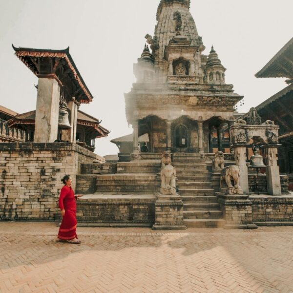 A woman in a red saree walks by an ancient temple with intricate architecture, stone sculptures of lions on stairs, and a traditional bell structure, in a square with herringbone-patterned brick flooring, basked in soft sunlight.