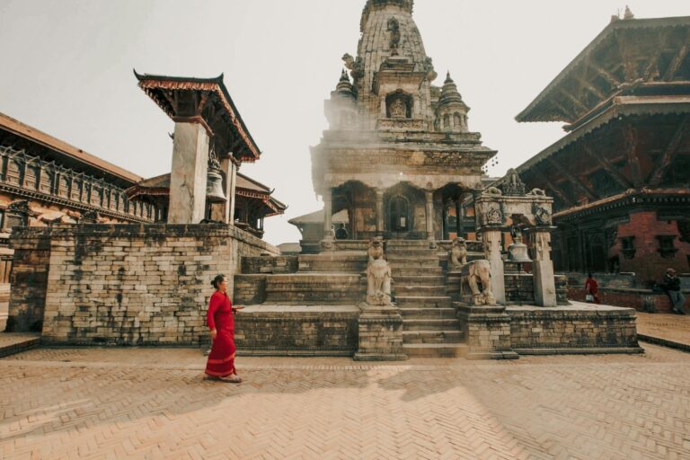 A woman in a red saree walks by an ancient temple with intricate architecture, stone sculptures of lions on stairs, and a traditional bell structure, in a square with herringbone-patterned brick flooring, basked in soft sunlight.