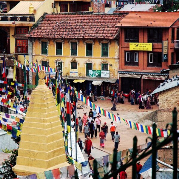 A busy street in Nepal with a golden stupa in the foreground, colorful prayer flags strung across, and traditional Nepalese architecture in the background. People are walking and gathering in the area.