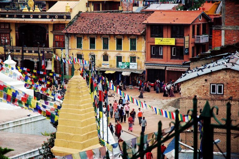 A busy street in Nepal with a golden stupa in the foreground, colorful prayer flags strung across, and traditional Nepalese architecture in the background. People are walking and gathering in the area.