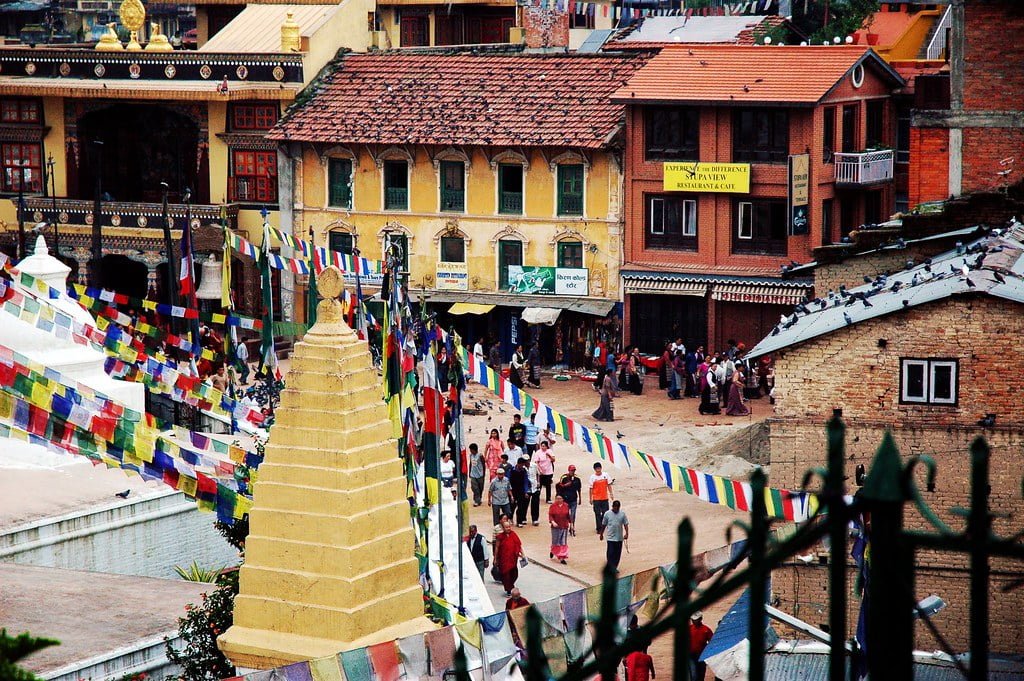 A busy street in Nepal with a golden stupa in the foreground, colorful prayer flags strung across, and traditional Nepalese architecture in the background. People are walking and gathering in the area.