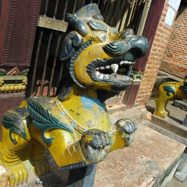 Two colorful statues of mythical lion-like creatures guard the entrance to a building, with weathered paint and intricate details, against a backdrop of a brick wall and traditional architecture.