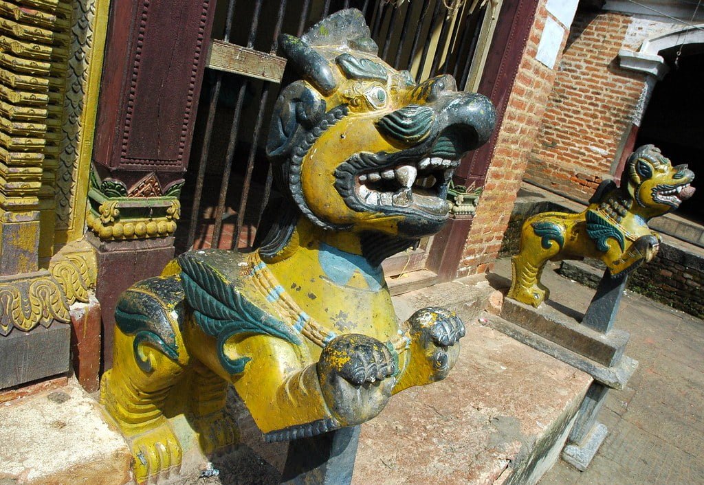 Two colorful statues of mythical lion-like creatures guard the entrance to a building, with weathered paint and intricate details, against a backdrop of a brick wall and traditional architecture.