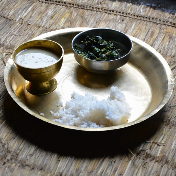 A traditional meal served on a brass plate with a heap of white rice, a bowl of green leafy vegetables, and a brass bowl of buttermilk, placed on a woven mat.