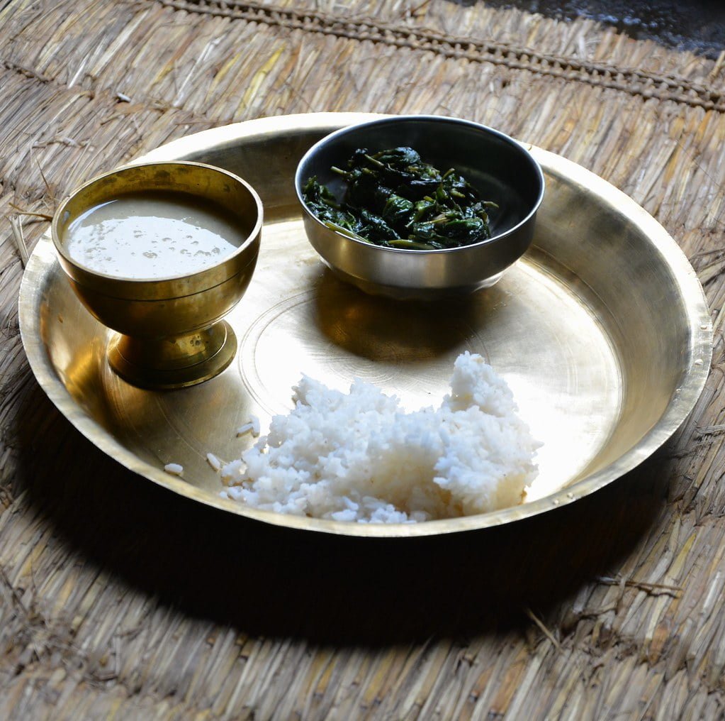 A traditional meal served on a brass plate with a heap of white rice, a bowl of green leafy vegetables, and a brass bowl of buttermilk, placed on a woven mat.