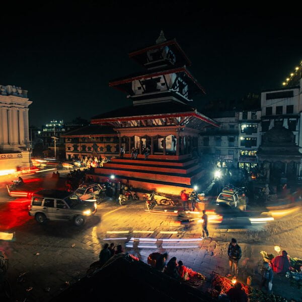 A bustling night scene at a city square with a traditional multi-tiered temple lit by ambient light, surrounded by motion-blurred vehicles and pedestrians, with some people gathered around vendors on the street.